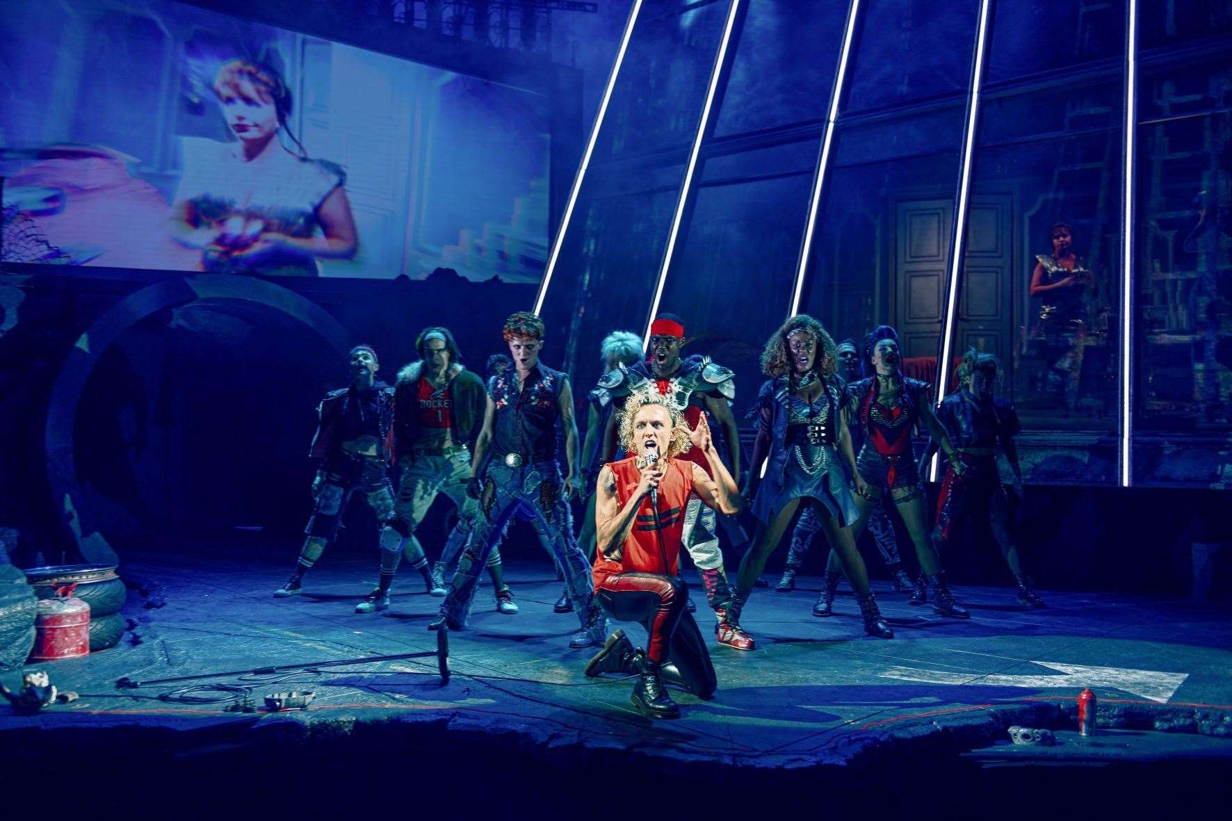 The musical has a unique staging with a main central area, a separate elevated section, and portions of the action are filmed live and projected onto video screens.