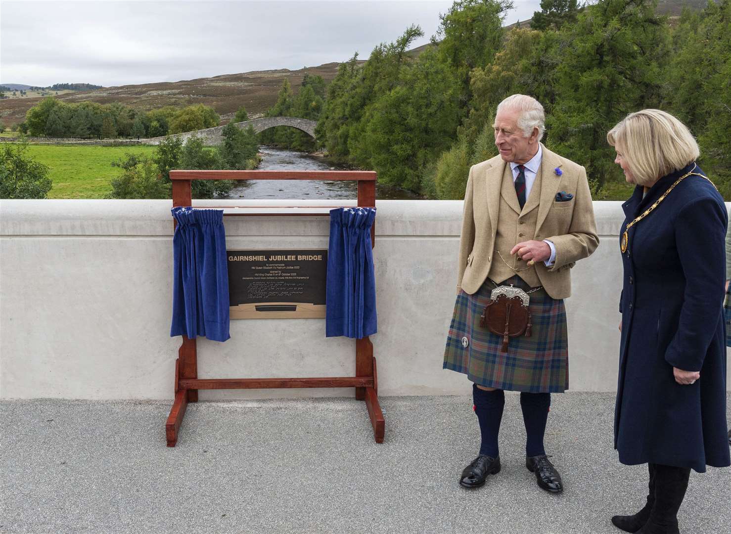 His Majesty The King unveils a plaque to formally name the Gairnshiel Jubilee Bridge watched by Provost of Aberdeenshire Cllr Judy Whyte